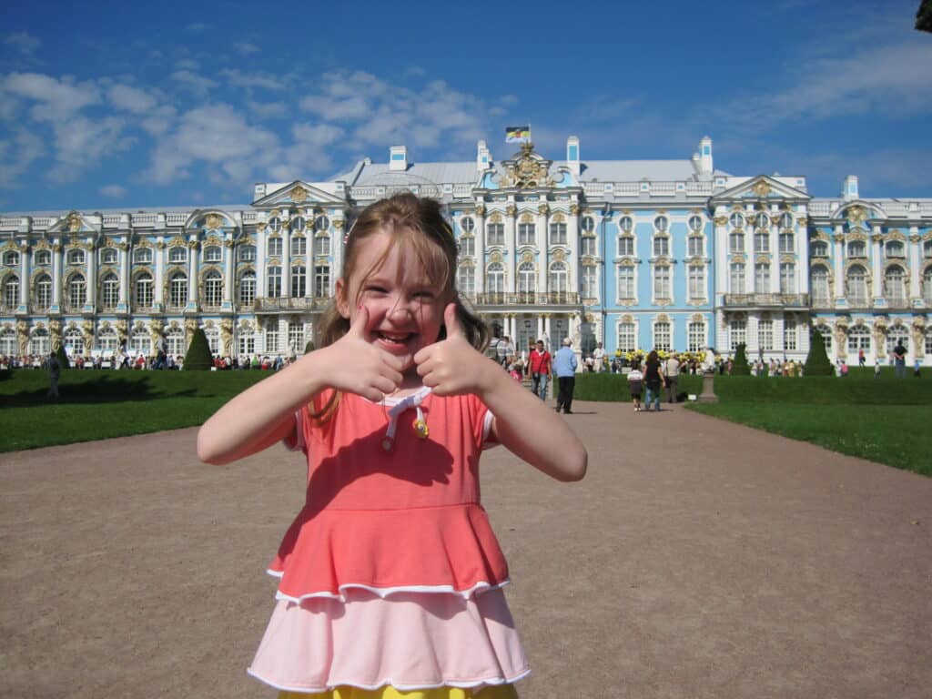 Smiling young girl in dress gives two thumbs up outside Catherine's Palace on a bright sunny day.