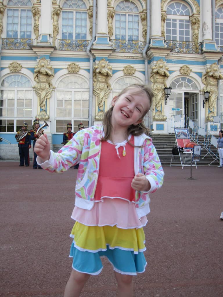 Smiling young girl dancing outside Catherine's Palace with uniformed band members playing in background.