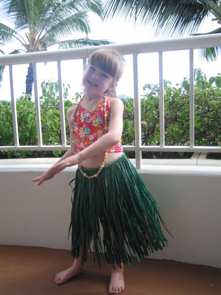 Young girl wearing pink flowered bathing suit top, white shell necklace and green grass skirt practicing hula dance on balcony at Fairmont Kea Lani in Maui.