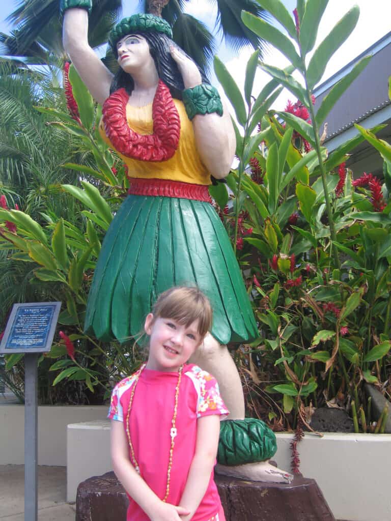 Young girl dressed in pink and wearing long chain necklace poses in front of sculpture of Hawaiian dancer.