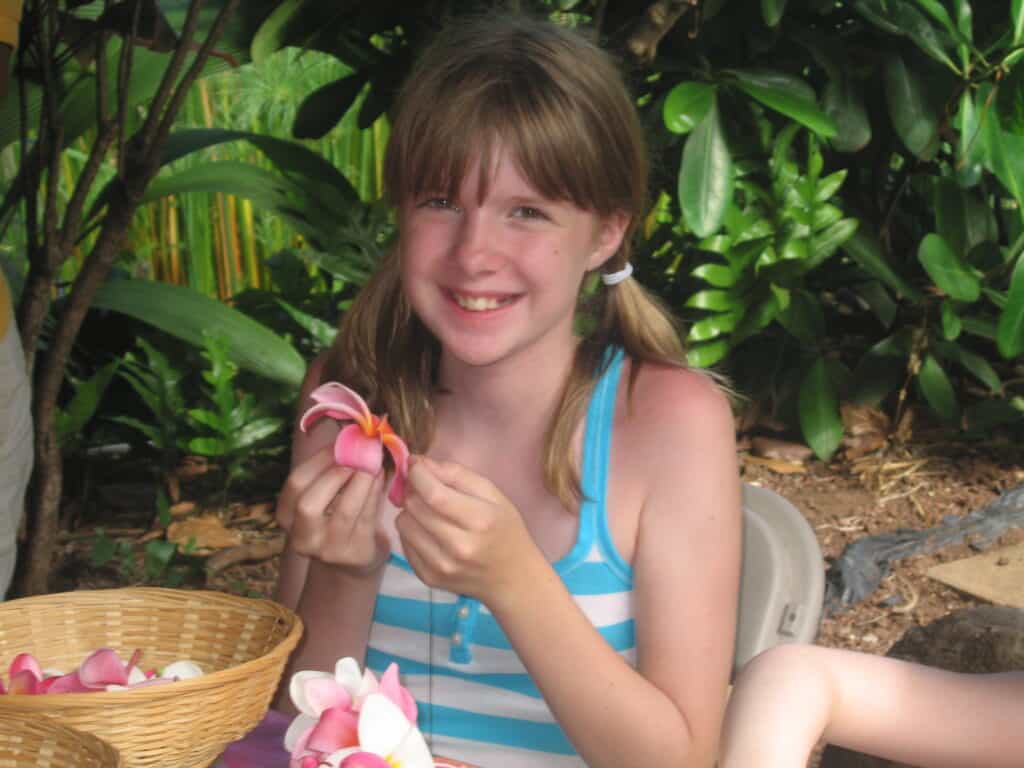 Young girl in blue and white striped tank top threading pink flower while making leis at Maui Tropical Plantation.