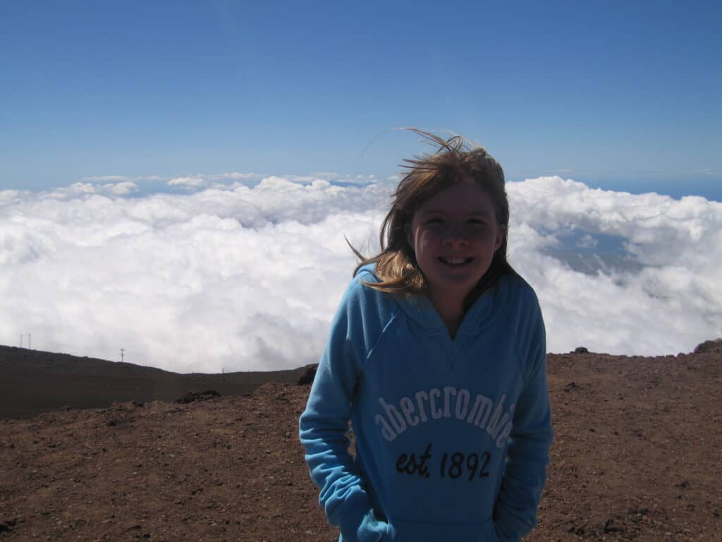 Young girl in blue sweatshirt posing at top of Haleakala Crater, Maui with clouds behind her.