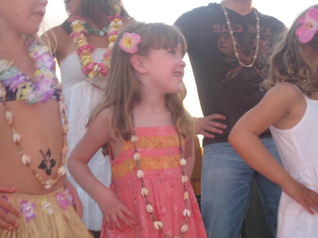 Young girl in coral dress wearing shell necklace and pink flower in hair learning hula in group at luau.