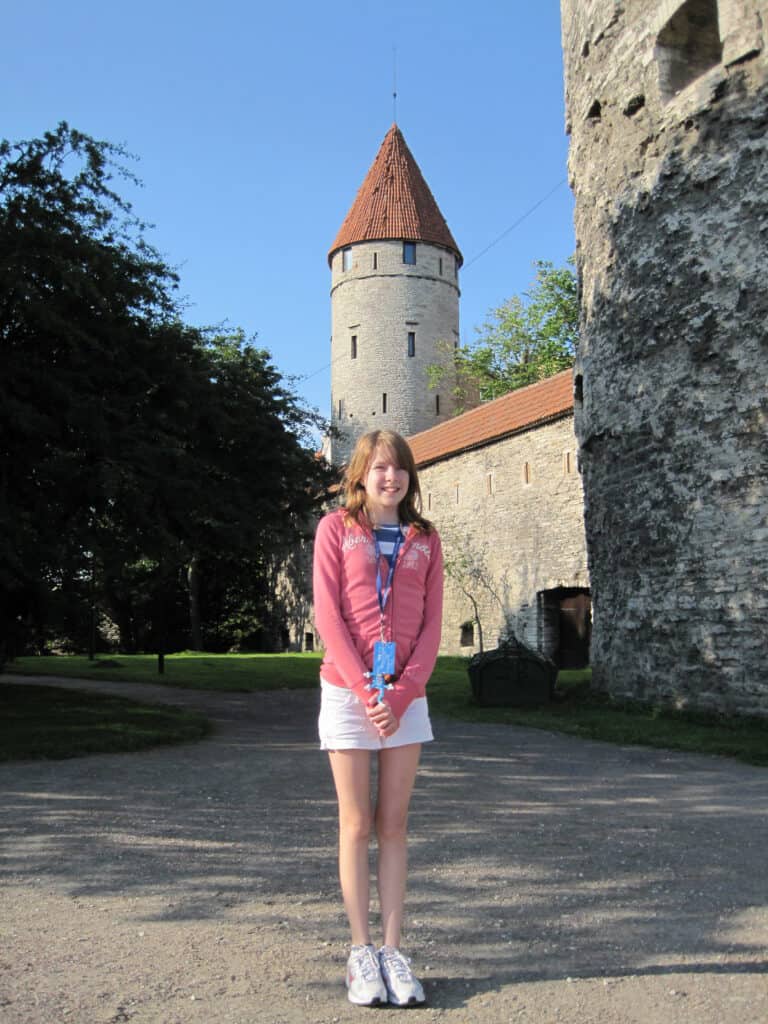 Young girl in pink sweatshirt and white shorts standing outside stone walls of Tallinn's Old Town with round stone tower with orange/red roof in background.