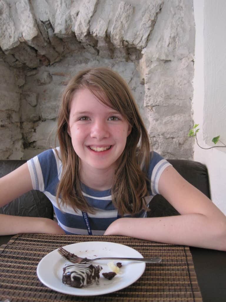 Young girl in blue striped shirt sitting at a cafe in Tallinn - stone walls behind her and woven placemat and white plate with fork and chocolate pastry with white piped icing in front of her.