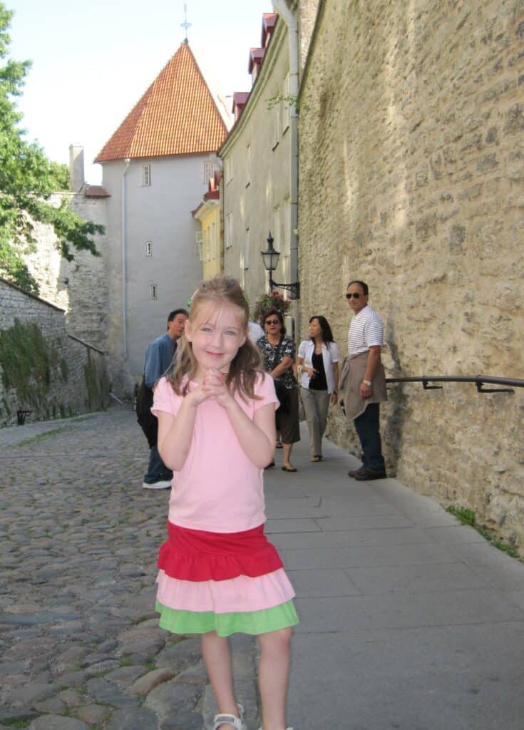 Young girl in pink t-shirt and red, pink and green ruffled skirt poses on cobblestoned street in Tallinn, Estonia with group of tourists in background.