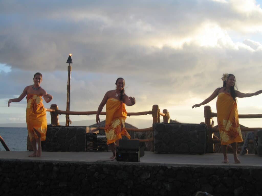 Three women in orange flowered dresses dancing on stage at sunset at luau in Maui.