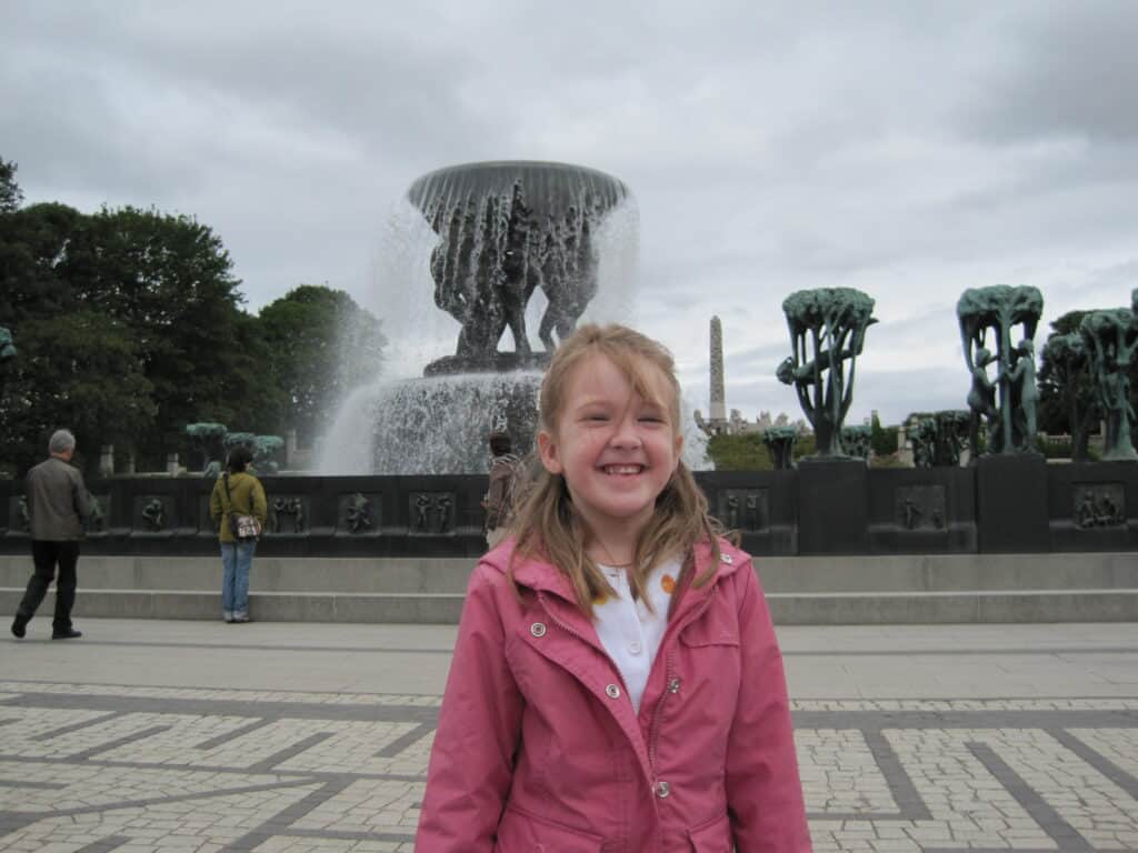 Smiling young girl in pink jacket standing in front of The Fountain at Vigeland Park in Oslo, Norway.