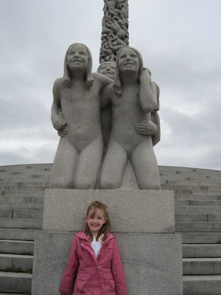 Young girl in pink jacket posing in front of two statues at the Monolith Plateau in Vigeland Park, Oslo, Norway.