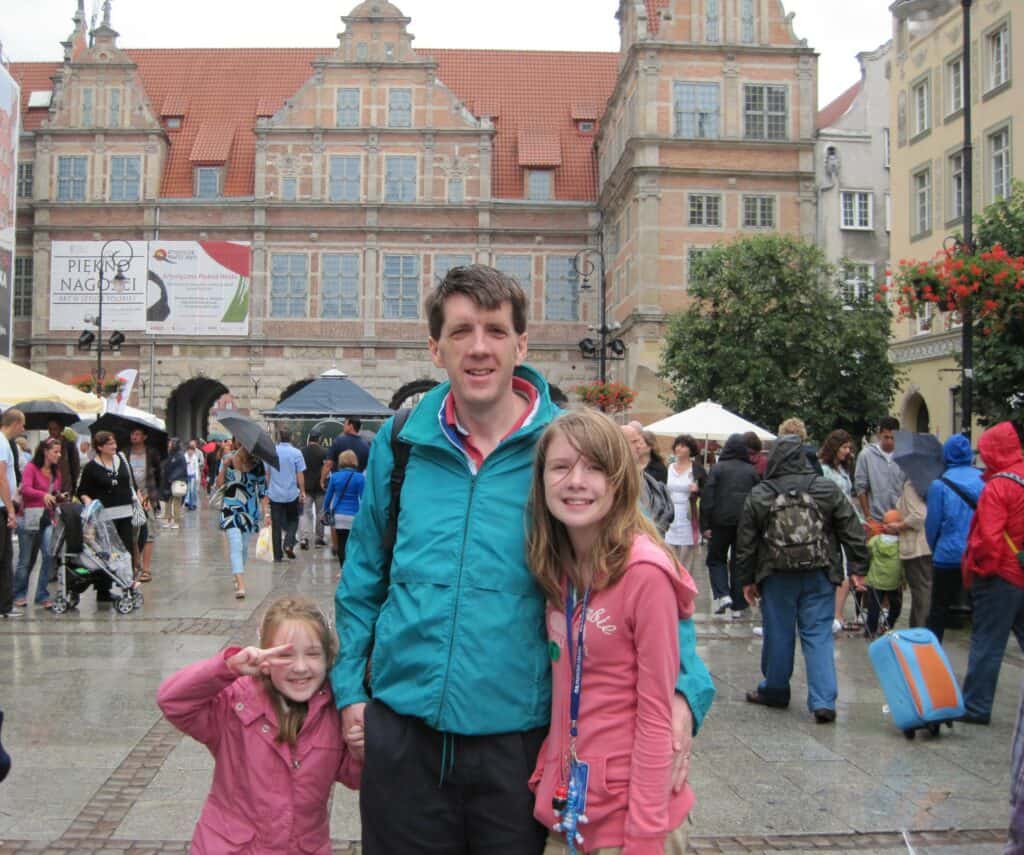 Dad in green rain jacket standing between younger and older daughters both in pink on wet, crowded street at the Green Gate in Gdansk, Poland.