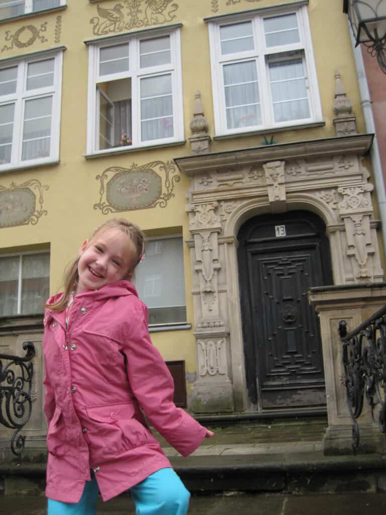 Young girl in pink rain coat posing in front of yellow building with elaborate door in Gdansk, Poland.