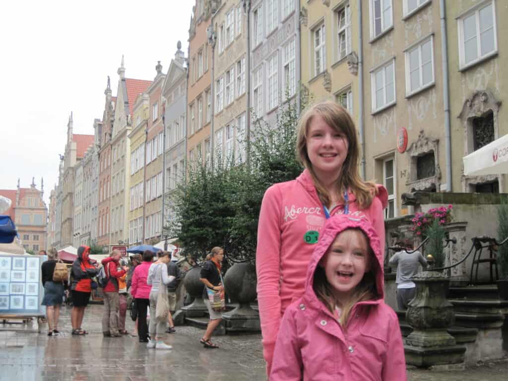 Older and younger girl dressed in pink standing on street in Gdansk on a rainy day with street market in background.