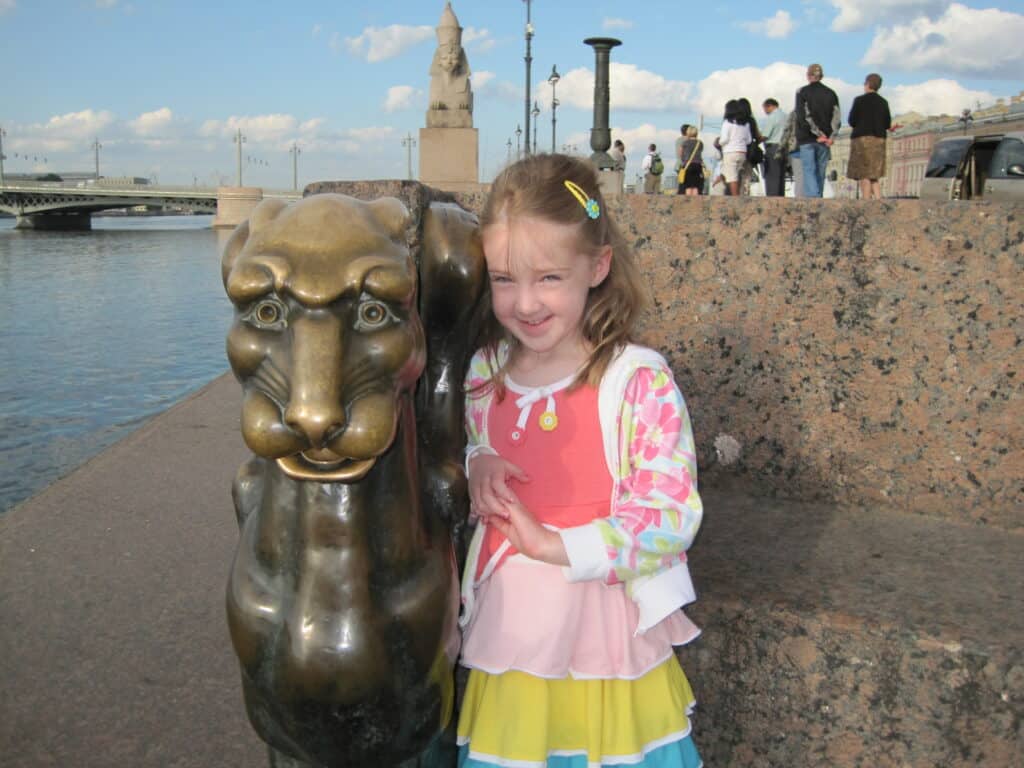 Young girl wearing dress and sweater standing beside bronze gryphon alongside Neva River in St. Petersburg, Russia.