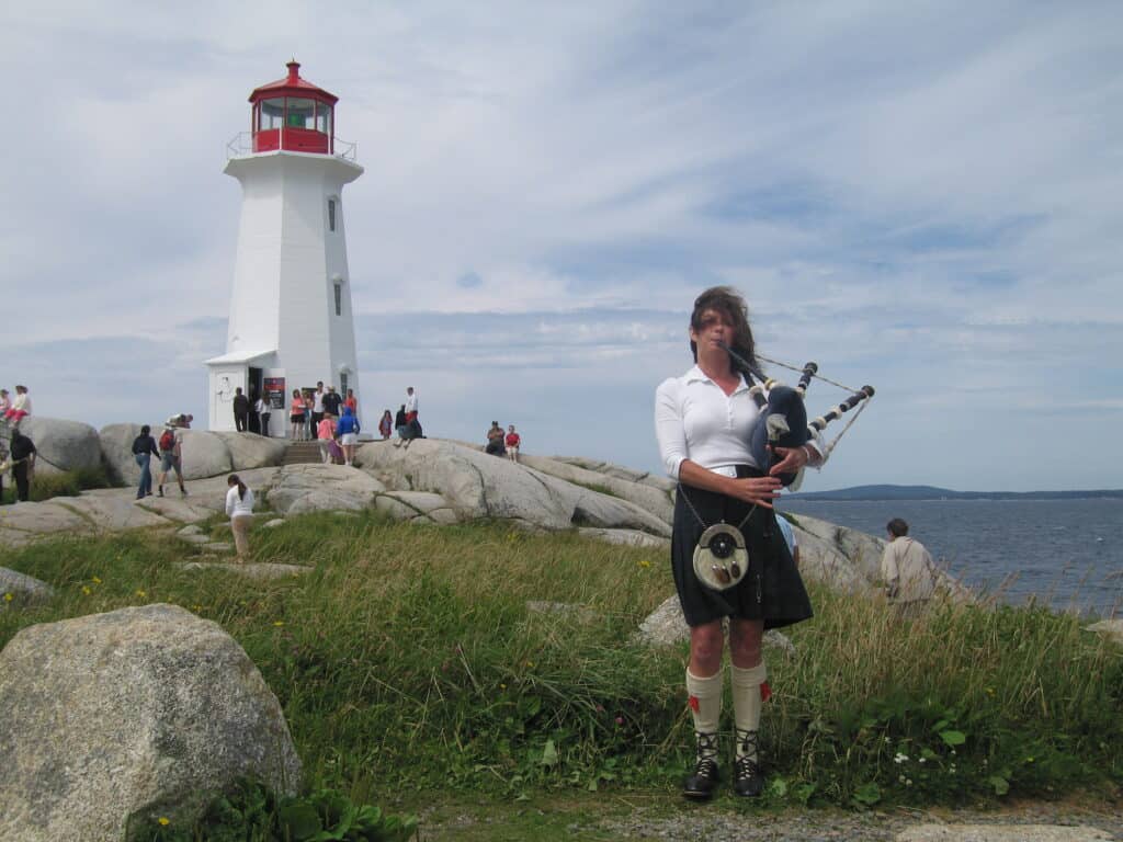 Woman in kilt and white top playing bagpipes with Peggy's Cove Lighthouse and ocean in background.