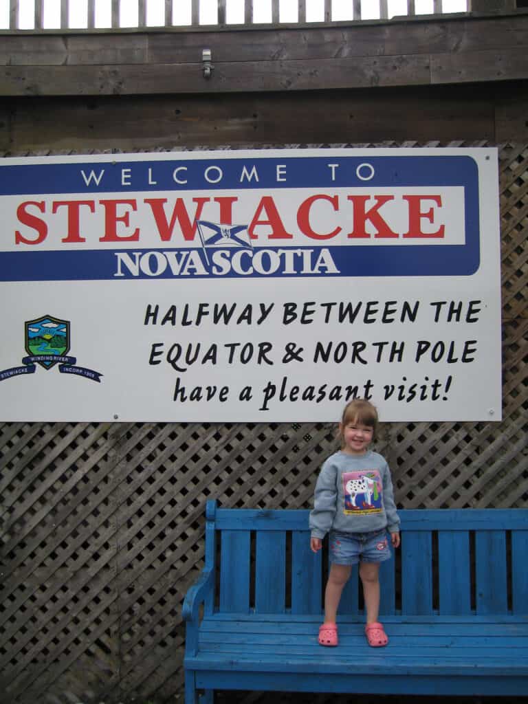 Young girl in shorts and sweatshirt stnding on blue bench under sign reading "Stewiacke, Nova Scotia - Halfway between the Equator & North Pole - have a pleasant visit!".