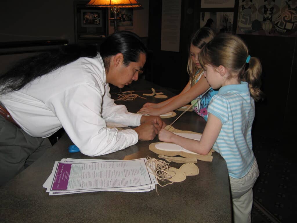 Native American man in white shirt helps young girl in blue stripped t-shirt thread leather through moccasins at Hyatt Regency, Scottsdale, Arizona.