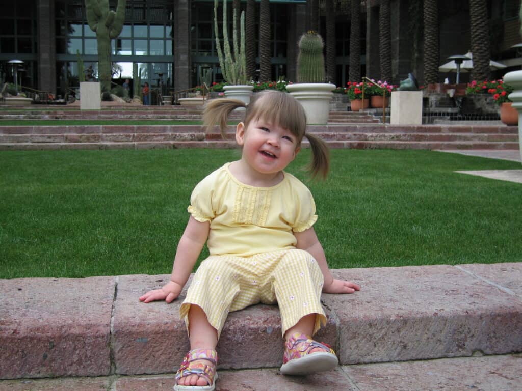 Toddler girl in pigtails dressed in yellow sitting on grounds of Hyatt Regency with terrace in background, Scottsdale, Arizona.