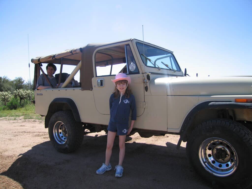 Young girl in navy blue shorts and long sleeved top and pink cowboy hat poses in front of cream-coloured jeep in Sonoran Desert.