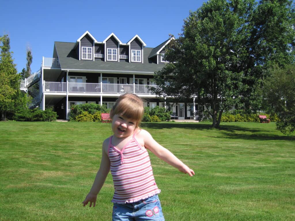 Young girl in striped tank top and denim shorts standing on lawn in front of building at Inverary Resort in Baddeck, Nova Scotia.