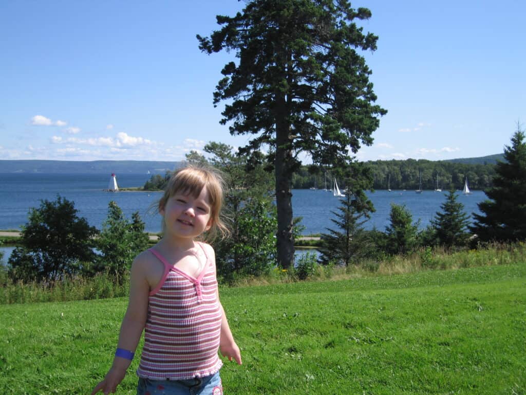 Toddler girl in striped tank top standing on grass outside Alexander Graham Bell National Historic Site with lake, sailboats and lighthouse in background.