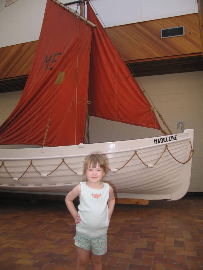 Young girl in tank top and shorts posing in front of Madeleine sailing boat (white boat with red sail) at the Maritime Museum of the Atlantic in Halifax, Nova Scotia.