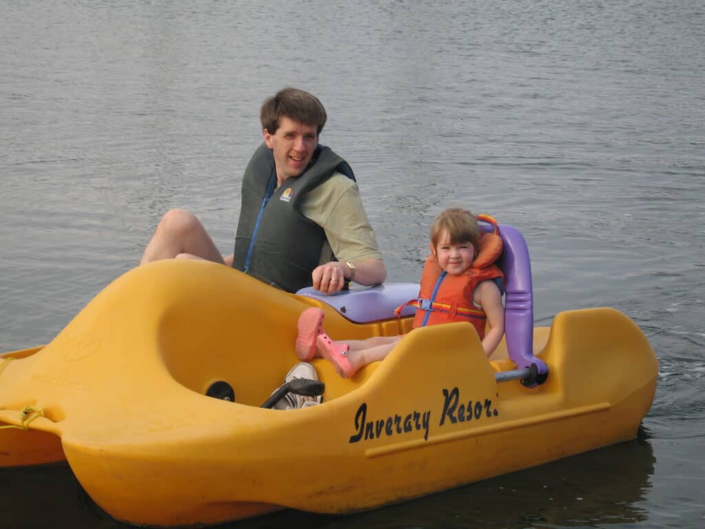 Man and toddler girl wearing life jackets in yellow pedal boat on lake at Inverary Resort, Nova Scotia.