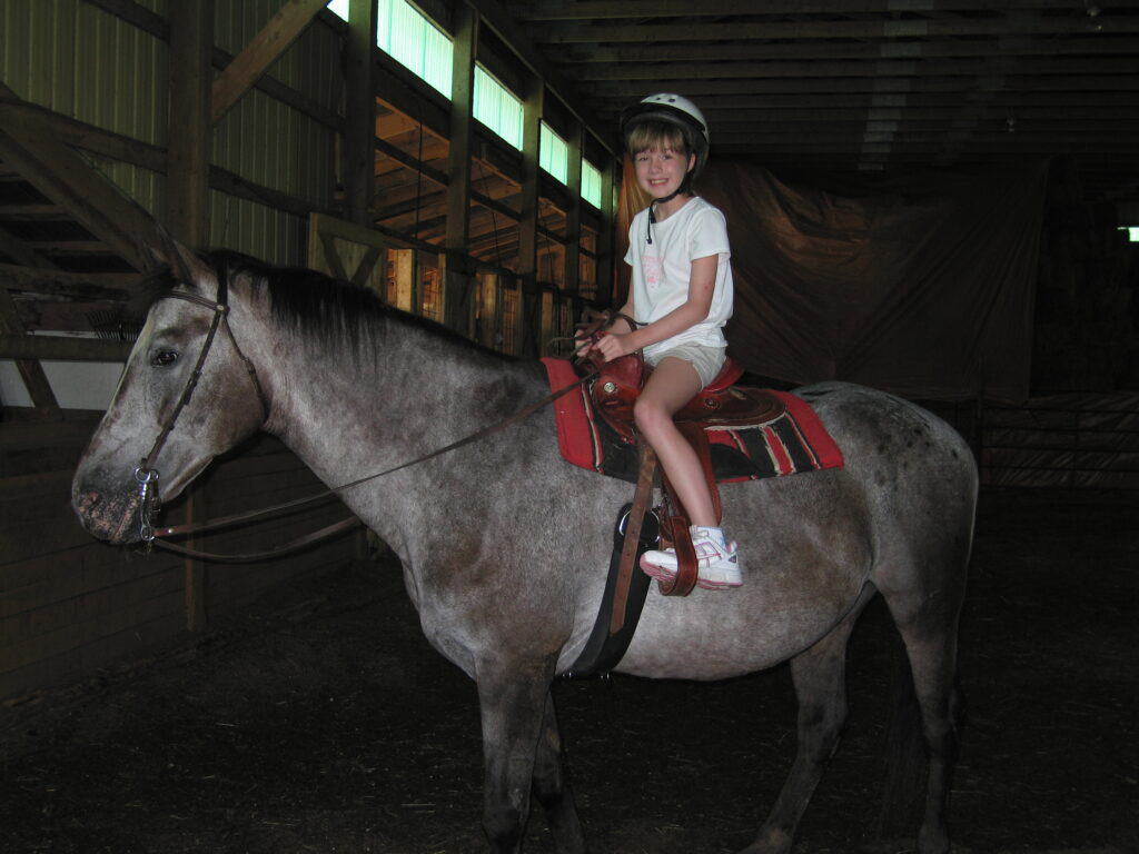 Young girl in shorts, t-shirt and helmet sitting on gray horse in stables in Baddeck on Cape Breton Island, Nova Scotia.