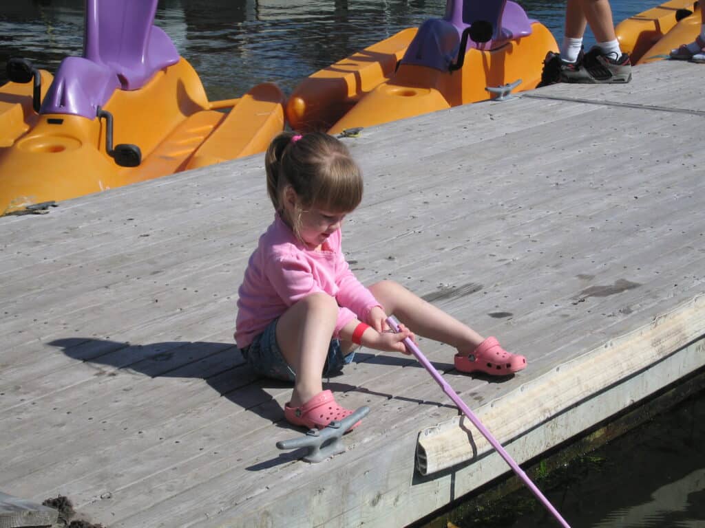 Toddler girl in pink short and crocs sitting on dock at Inverary Resort holding pole into the water with yellow and purple pedal boats in background.