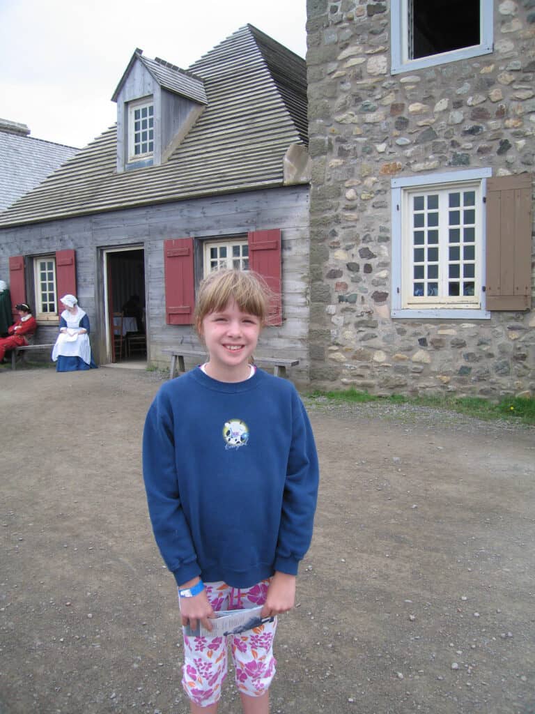 Young girl in dark blue sweatshirt and pink flowered pants standing in front of stone buildings at the Fortress of Louisbourg, Cape Breton Island.