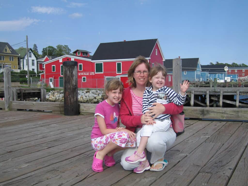 Woman and two young girls on dock in Lunenburg with colourful buildings in background.