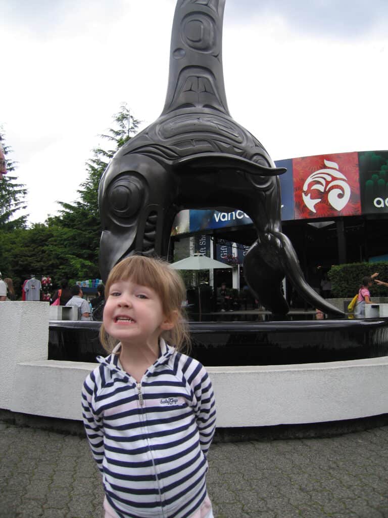 Young girl in striped hoodie poses in front of sculpture outside Vancouver Aquarium.