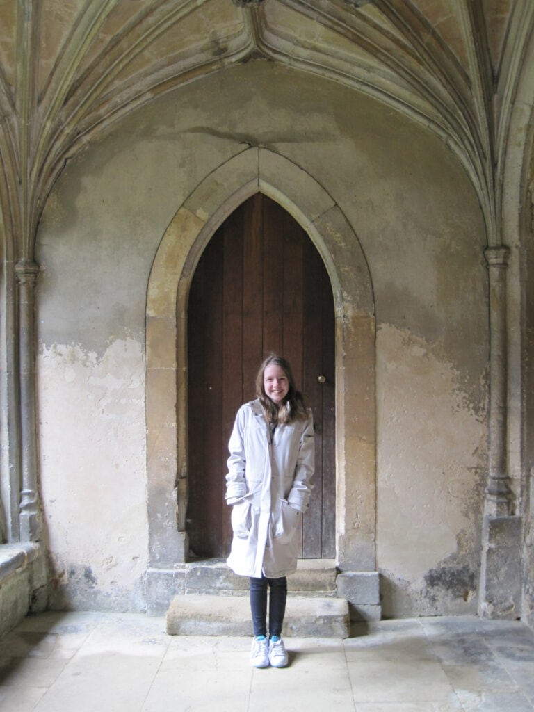 young teen girl in black pants and light coloured rain coat standing in Lacock Abbey on Harry Potter tour.