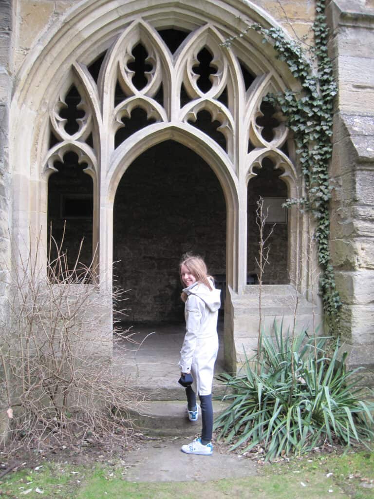 young girl in dark pants and light coloured coat turns to look back as she walks through stone archway at Oxford University.