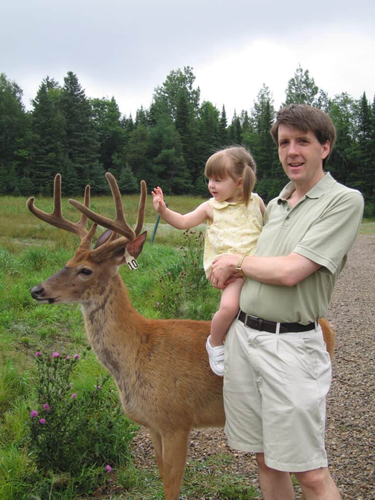 man holding toddler girl reaching out to touch antlers of a deer at Parc Omega in Montebello, Quebec.