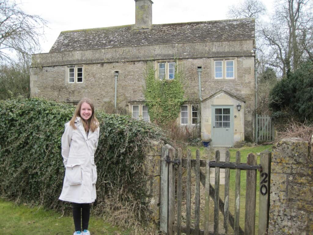 Young girl standing by ivy-covered fence and front gate with stone house in background on Harry Potter tour of Lacock, England.