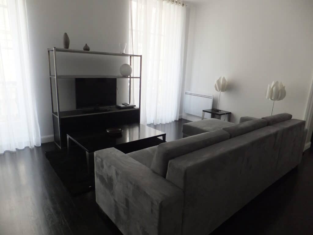 Living area of apartment in Paris with white walls, dark wood floors, white sheer curtains on windows, grey sofa and dark coffee table, metal tv stand with small television and three small vases.