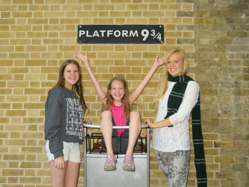 tween girl in pink top sitting in luggage cart with hands in air and a teenage girl standing on either side in front of brick wall and Platform 9 3/4 sign at King's Cross station in London.