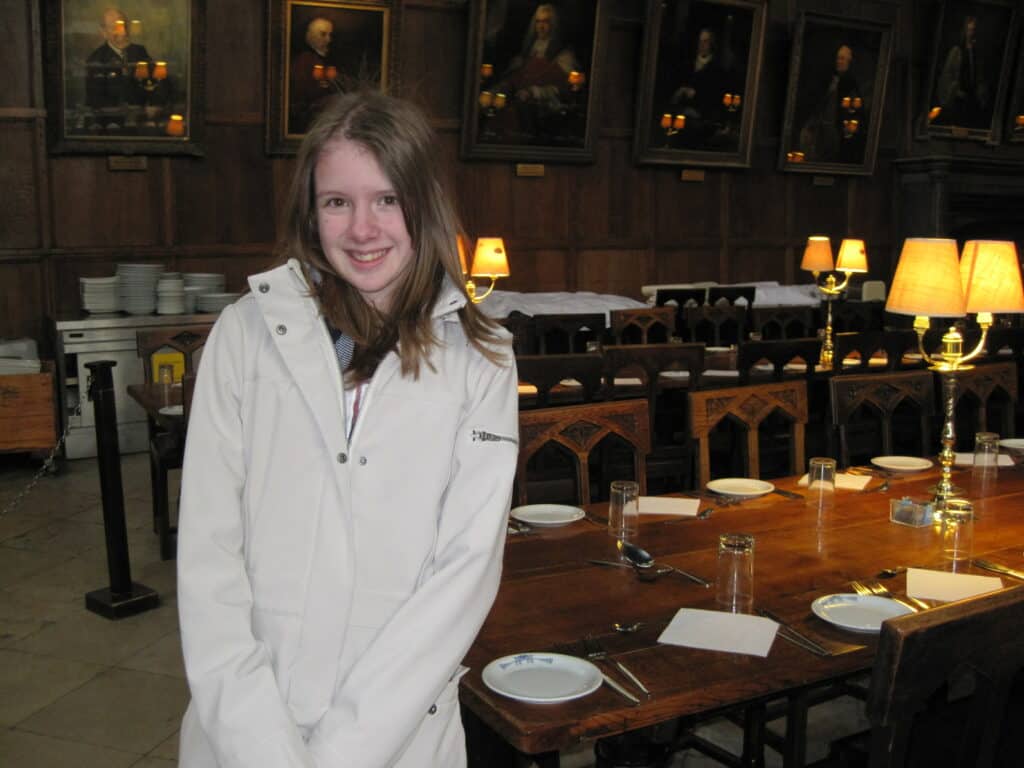 young girl in light coloured coat standing by table set with plates, glasses, napkins, cutlery and lit by lamps in dining hall at oxford university.