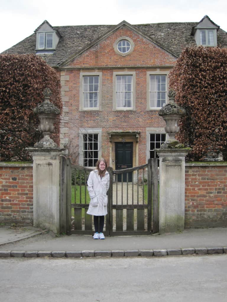 Young girl standing beside brick and stone front gate and wall with red brick home in background on Harry Potter tour of Lacock.