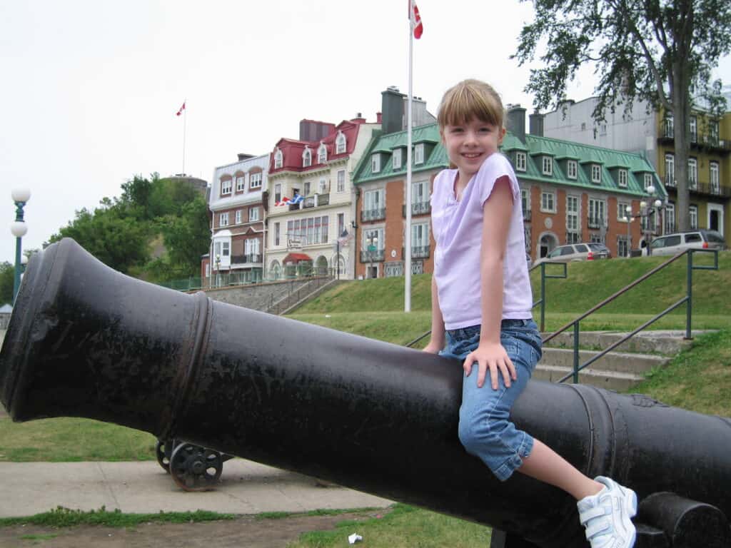 Young girl wearing capri jeans and light purple t-shirt sits on cannon on Terrasse Dufferin with buildings in background in Quebec City, Canada.