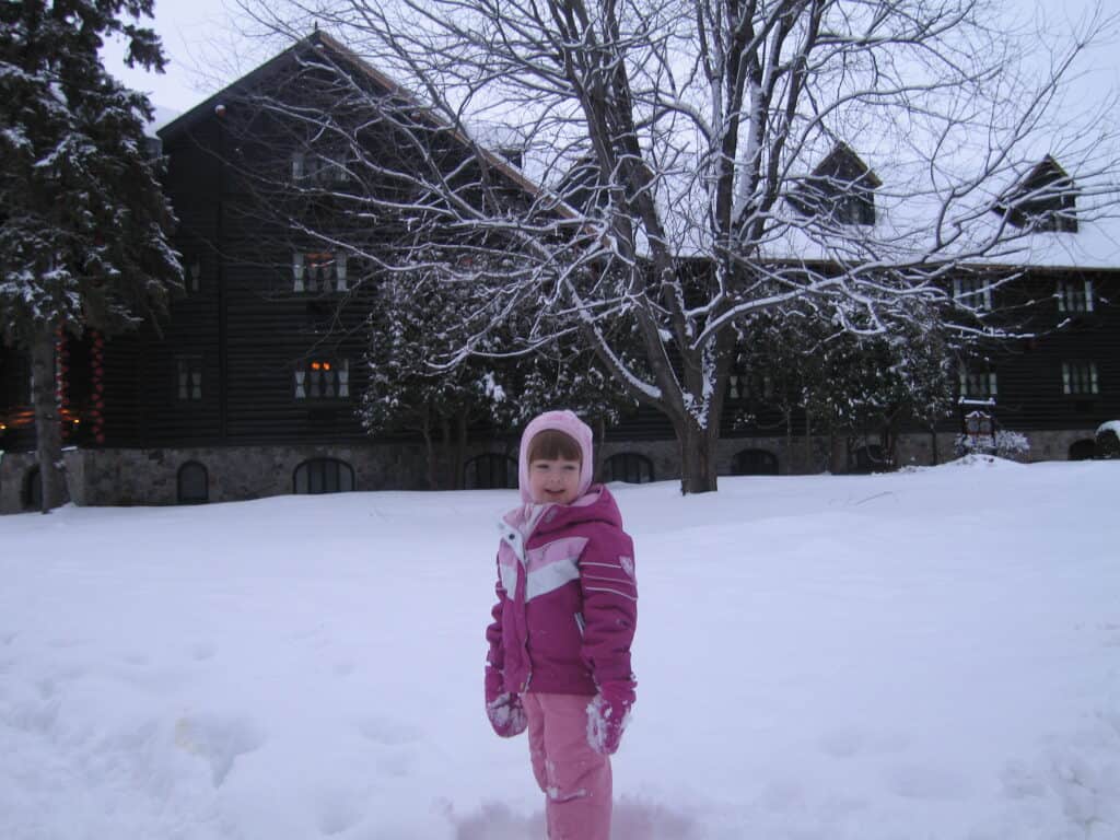 Young girl in pink snowsuit and hat standing in snow in front of Chateau Montebello.