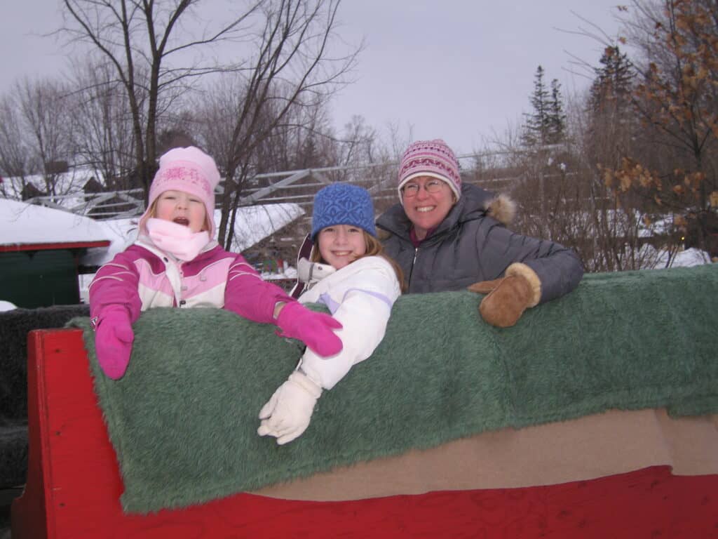 mom and two daughters in back of red wooden sleigh with green blanket.