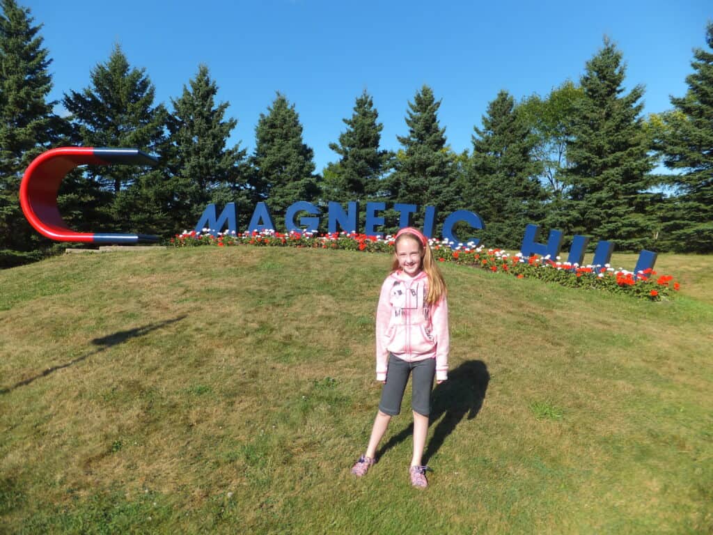 Young girl in pink sweatshirt and long grey shorts stands on grass in front of Magnetic Hill sign in New Brunswick.