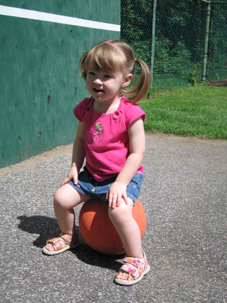 toddler girl in denim shorts and pink tshirt sitting on basketball on outdoor court at chateau montebello.