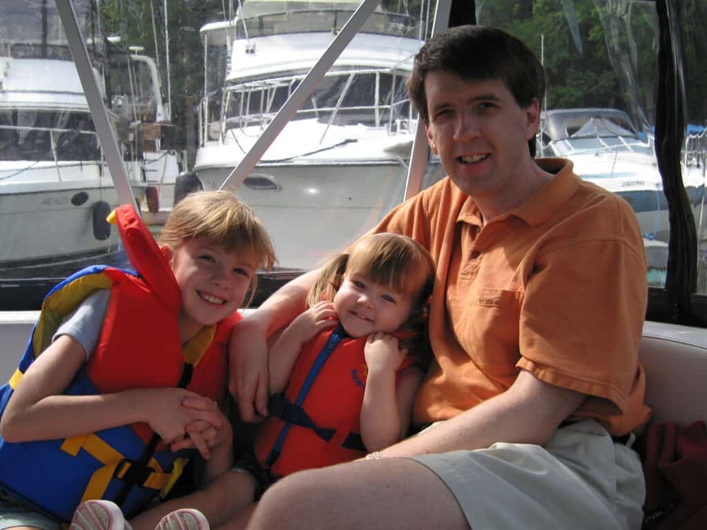 Two young girls wearing life jackets sit with dad in boat at Chateau Montebello with other boats in background.