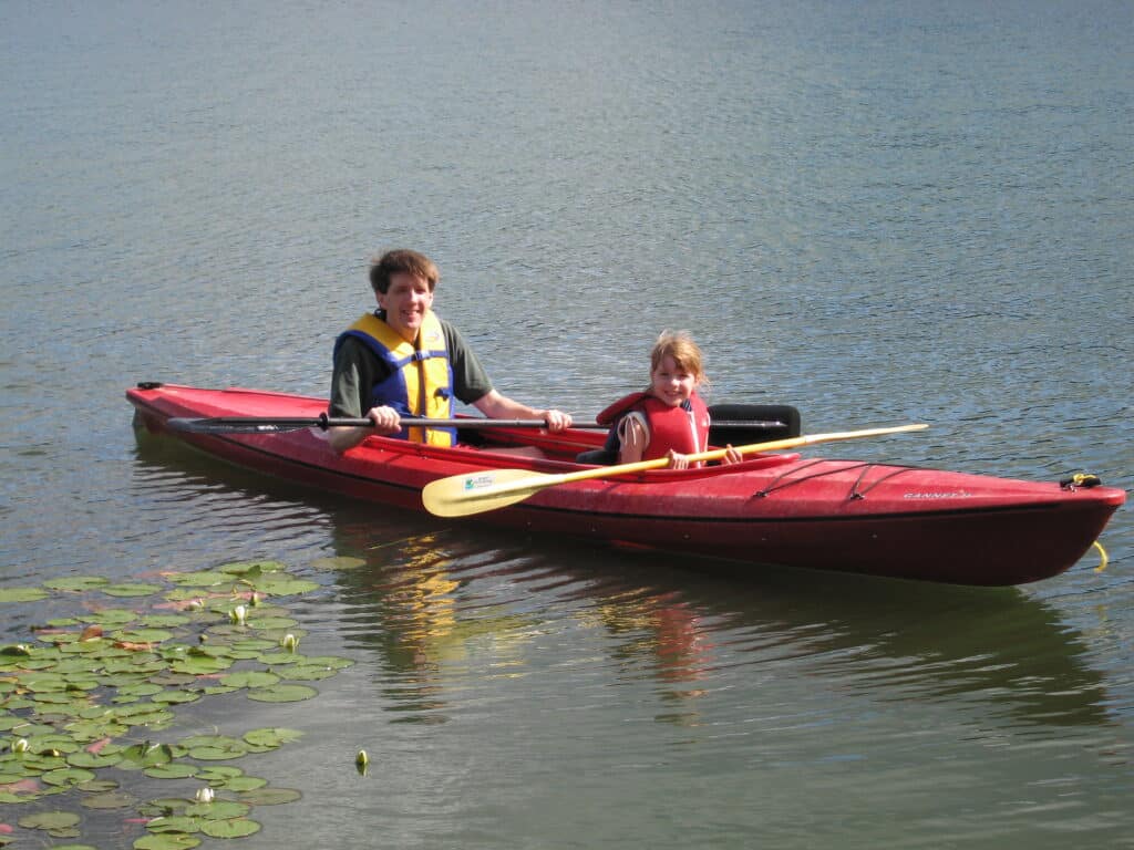 Dad and daughter wearing lifejackets in red kayak on lake with some lily pads to the left of the kayak at Whitefish Lake in Montebello, Quebec.