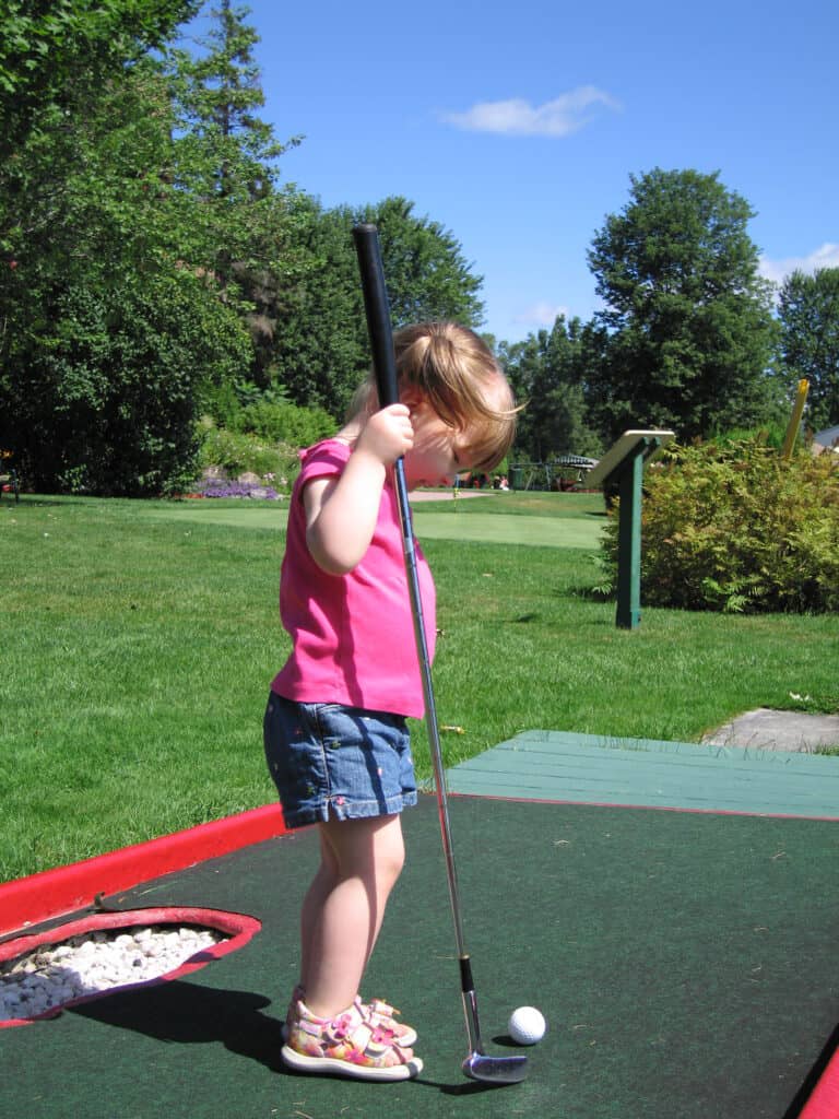 young toddler girl in denim shorts and pink t-shirt holding golf club and looking at golf ball on mini-putt course at chateau montebello.