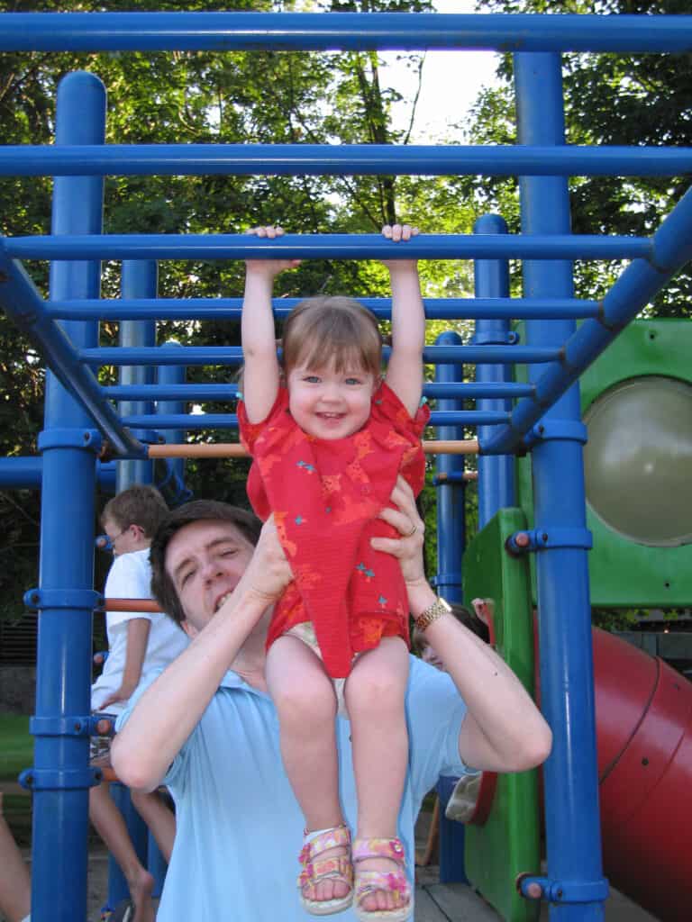 Man holding toddler girl in red dress as she hangs from bright blue bars at the Fairmont Chateau Montebello playground.