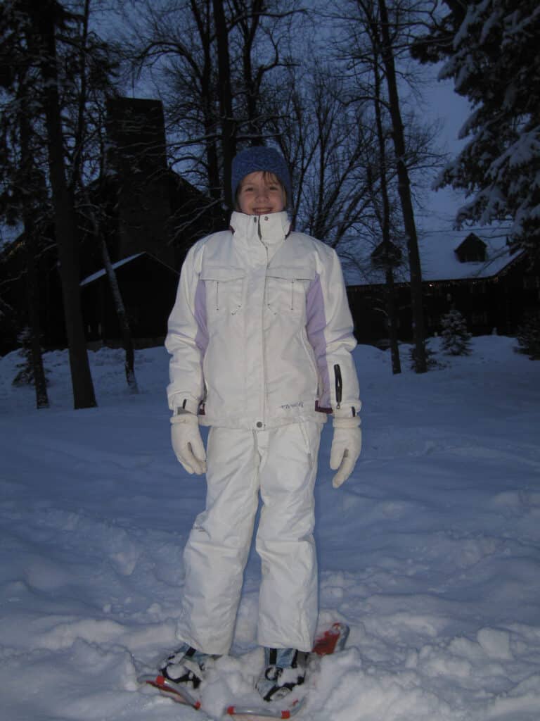 young girl in white snow pants and jacket on snowshoes at dusk with chateau montebello in background.