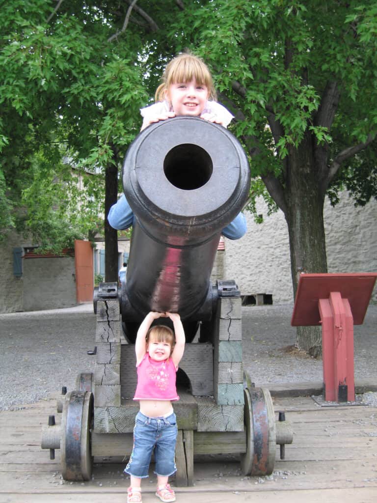 Two young girls posing with cannon in lower town of Quebec City - one sitting on cannon and one standing under.